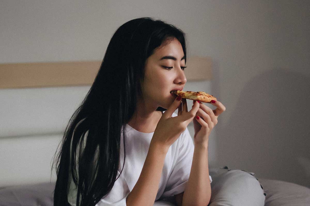 young woman eating a slice of pizza in bed