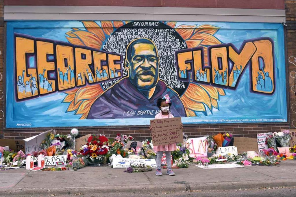 a young black girl holds a sign in front of a George Floyd mural